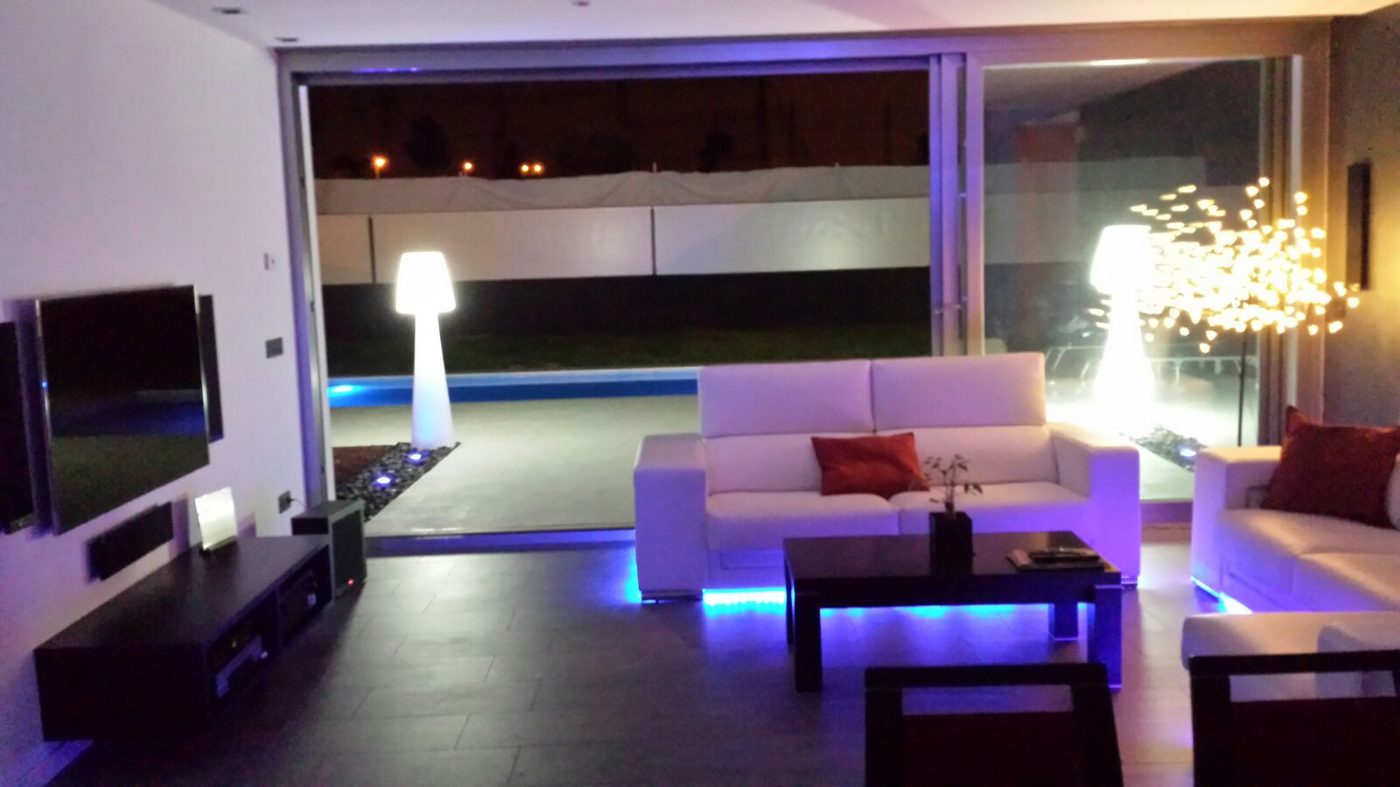 Salón abierto al porche en una vivienda terminada, con iluminación LED de color y la piscina iluminada al fondo